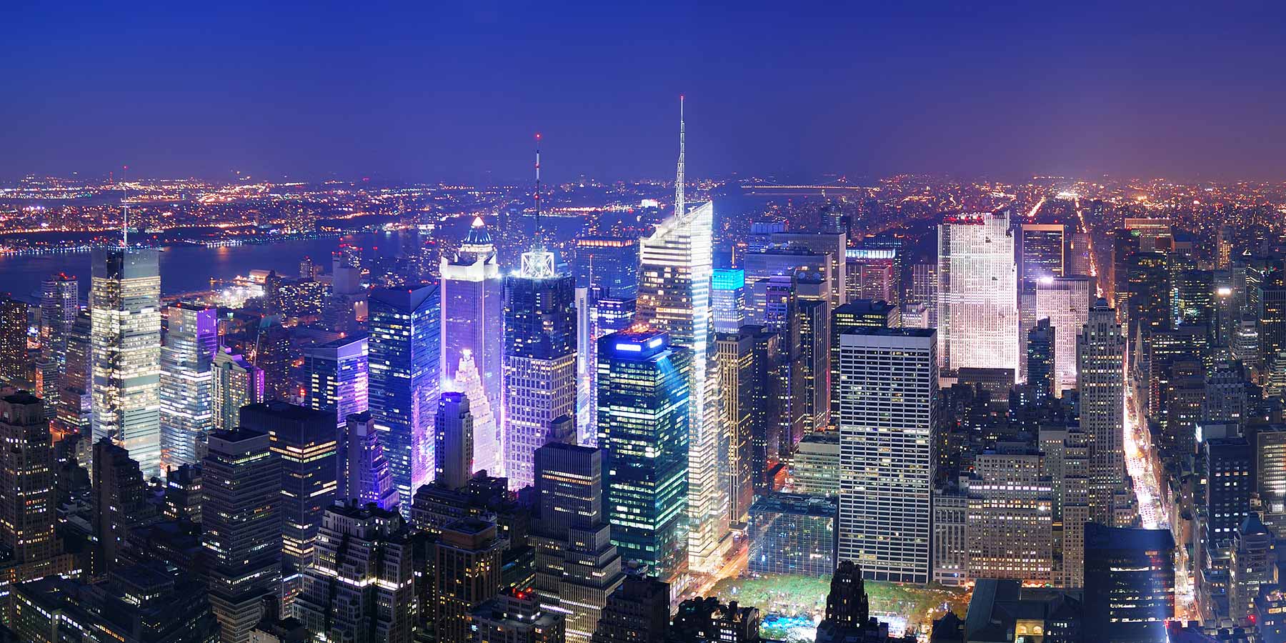 Aerial view of a brightly lit cityscape at night, with tall skyscrapers and a river visible in the background under a dark blue sky.